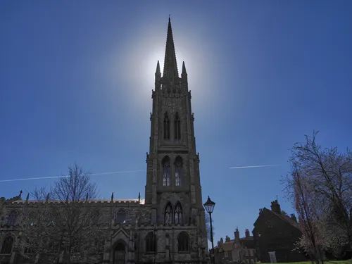 This architectural photograph captures St James' Spire, the prominent tower of St James' Church in Louth, Lincolnshire, positioned against a bright midday sky. It is springtime, as evidenced by the budding trees and clear conditions, which complement the church's stone façade. The urban setting is highlighted by surrounding historic buildings, typical of Louth, and there is a noticeable contrail in the sky, adding a dynamic element to the composition. The image was taken in the afternoon, showcasing the church as a significant landmark and example of urban architecture in Lincolnshire during the spring season.